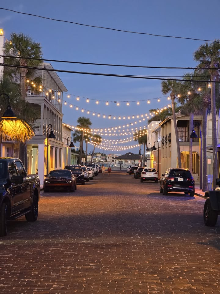 Cobblestone street lined with parked cars and palm trees, decorated with string lights at dusk.